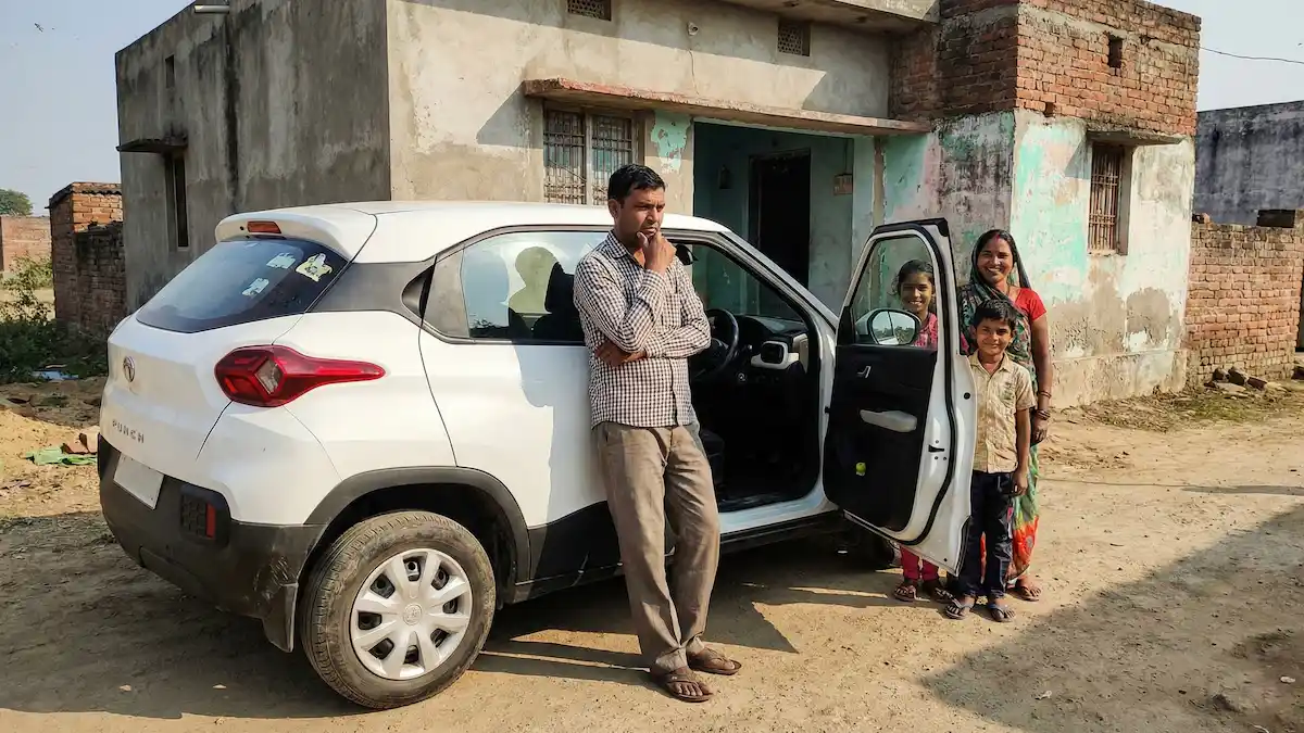 एक भारतीय परिवार अपनी नई टाटा पंच के साथ खड़ा है, बैकग्राउंड में गांव का घर या शहरी सोसायटी, सुरक्षित महसूस करते हुए।